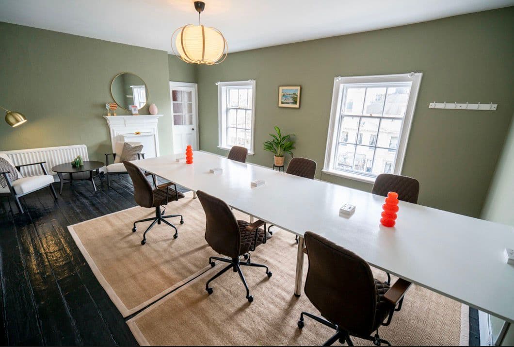 A meeting room with a long white table, green walls and seating for eight